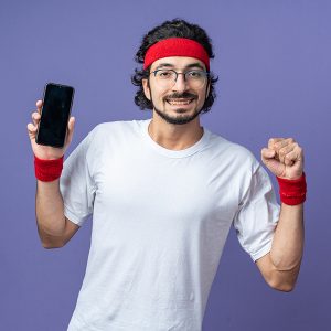 smiling young sporty man wearing headband with wristband holding phone showing yes gesture isolated on blue background